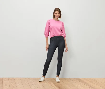 Une femme se tient sur un plancher en bois, portant un chemisier rose et un jean bleu foncé.