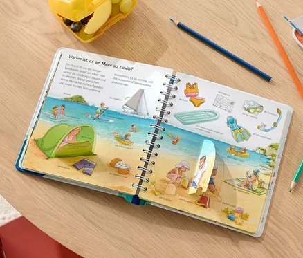 Un livre ouvert « Pourquoi ? Comment ? Junior : Nous partons en vacances » montre des illustrations d'une plage avec des personnes et des accessoires de plage.