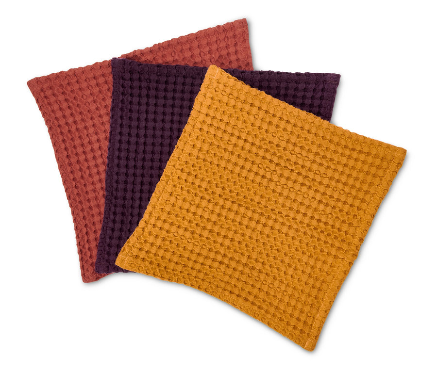 Trois serviettes de table en tissu texturé de couleurs orange, violet et rouge-brun posées sur un fond blanc.