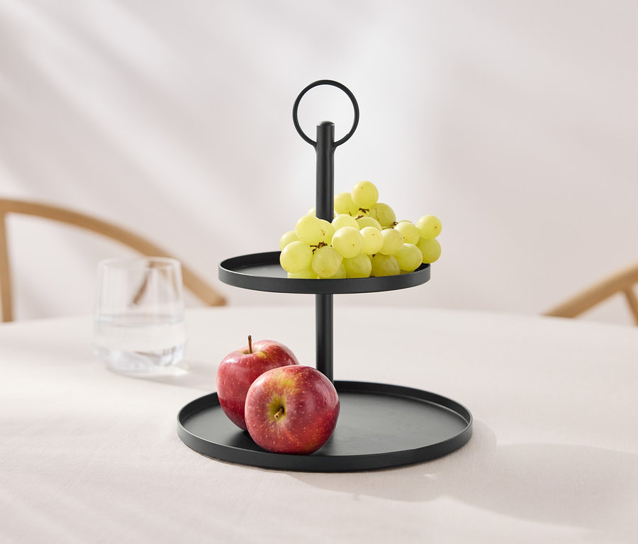 Un présentoir à deux étages noir avec des raisins et des pommes sur une table.