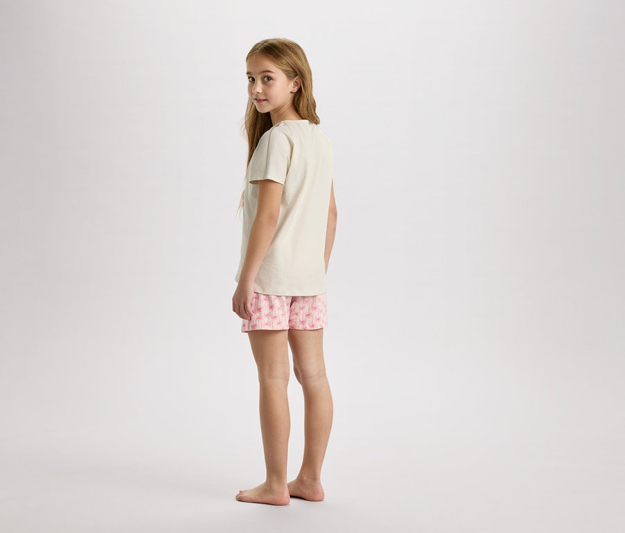Une jeune fille se tient pieds nus sur un fond neutre, portant un t-shirt beige et un short rose à imprimé floral.
