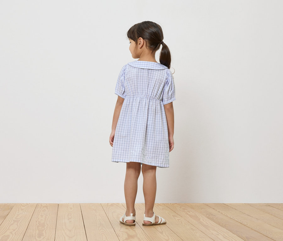 Une jeune fille, de dos, porte une robe à carreaux bleu et blanc et des sandales.