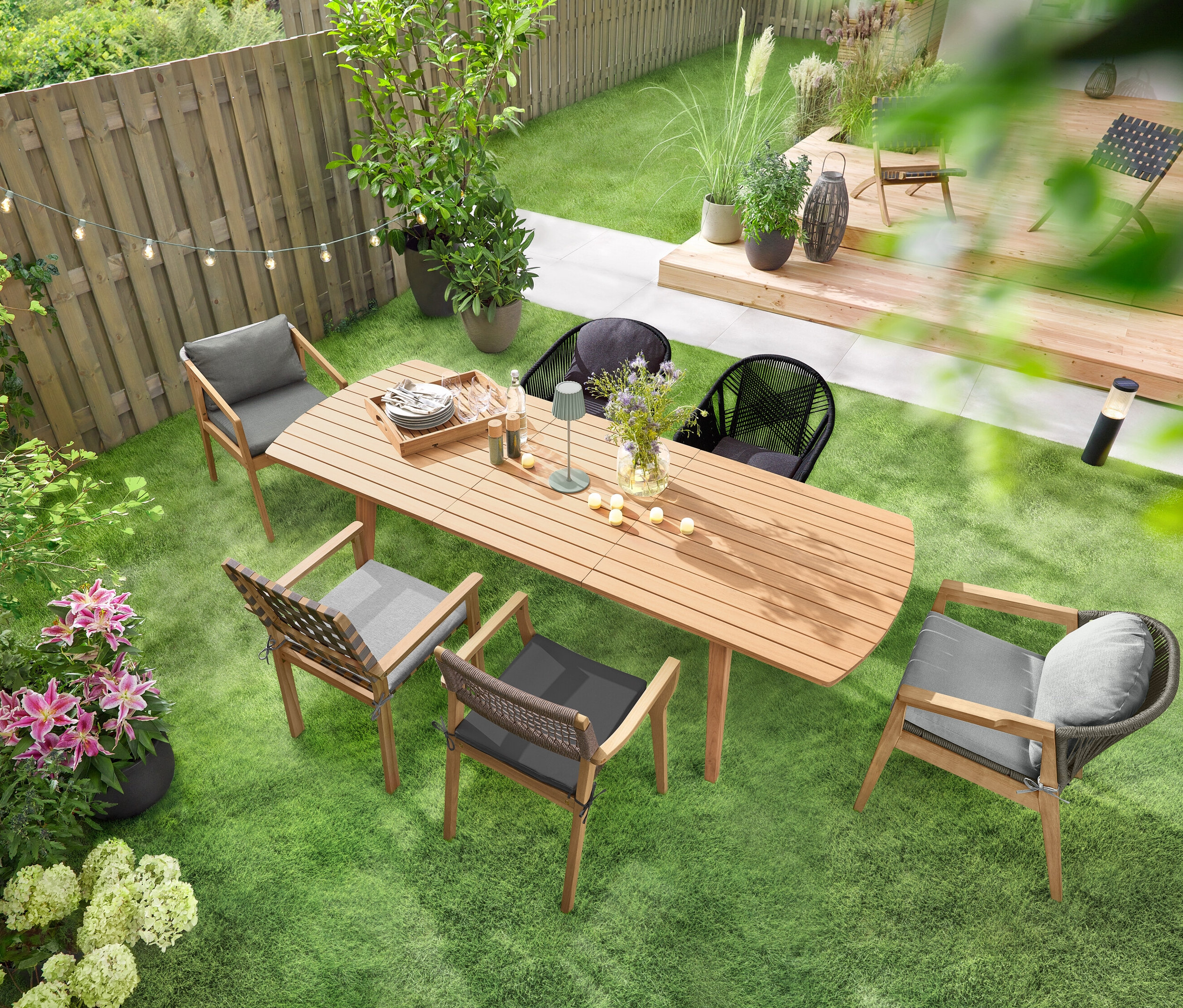 Une table de jardin à rallonges avec façades arrondies est posée sur une pelouse verte, entourée de plusieurs chaises de jardin. Une guirlande lumineuse solaire « Ampoules » est suspendue au-dessus de la table, et une lampe de table d’extérieur à LED rechargeable sauge est posée dessus. Plusieurs 6 bougies LED rechargeables d’extérieur sont réparties sur la table.