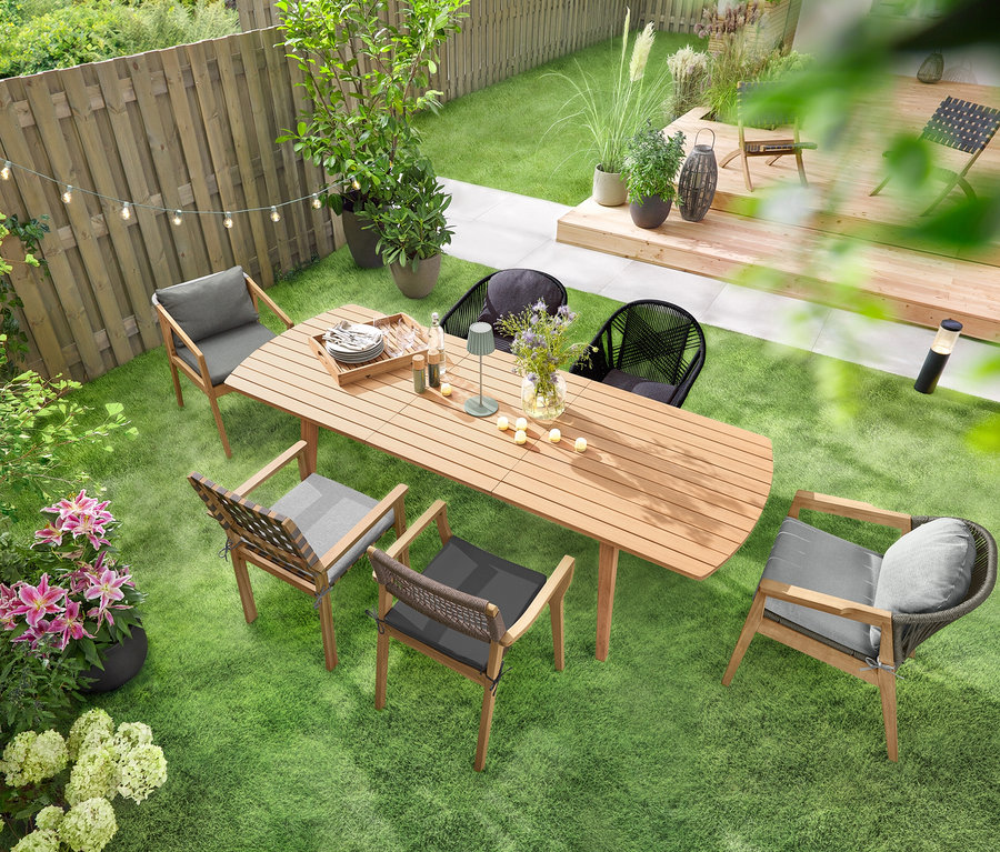 Une table de jardin à rallonges avec façades arrondies est posée sur une pelouse verte, entourée de plusieurs chaises de jardin. Une guirlande lumineuse solaire « Ampoules » est suspendue au-dessus de la table, et une lampe de table d’extérieur à LED rechargeable sauge est posée dessus. Plusieurs 6 bougies LED rechargeables d’extérieur sont réparties sur la table.