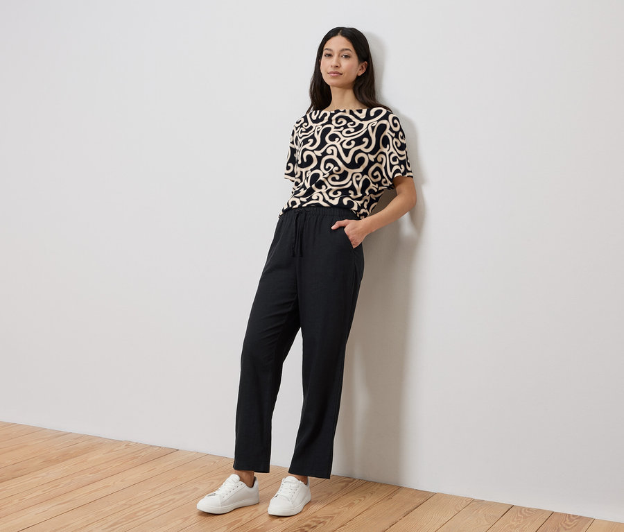 Femme appuyée contre un mur portant un haut à motifs, un pantalon noir et des baskets blanches.