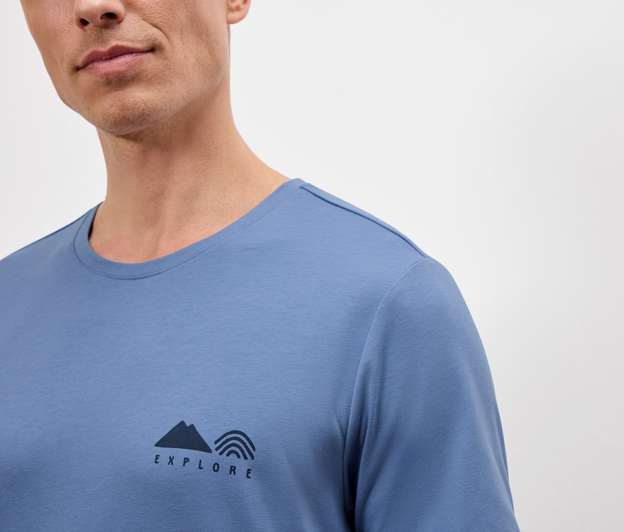Un homme porte un t-shirt bleu avec l'inscription "Explore" comprenant un symbole de montagne et un arc-en-ciel.