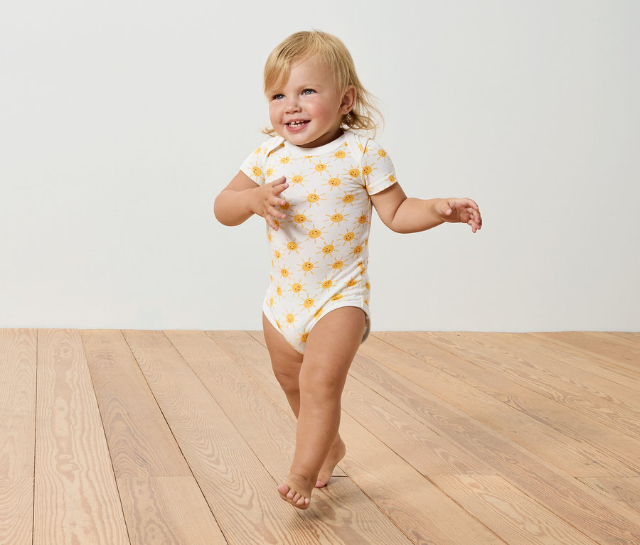 Un bébé blond avec un body à motifs de soleil marche sur un plancher en bois.