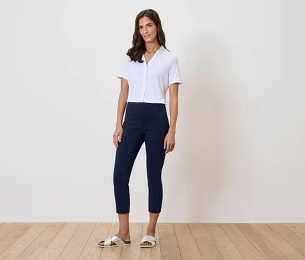 Une femme se tient devant un mur, portant une chemise blanche, un pantalon bleu et des sandales dorées.