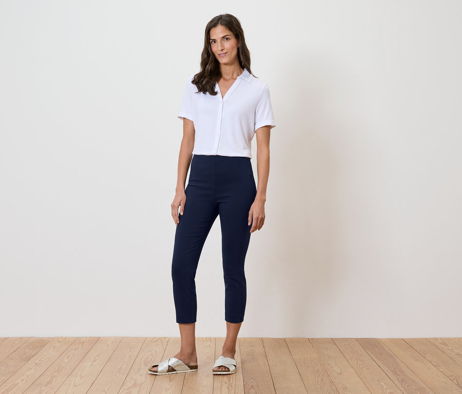 Une femme se tient devant un mur, portant une chemise blanche, un pantalon bleu et des sandales dorées.