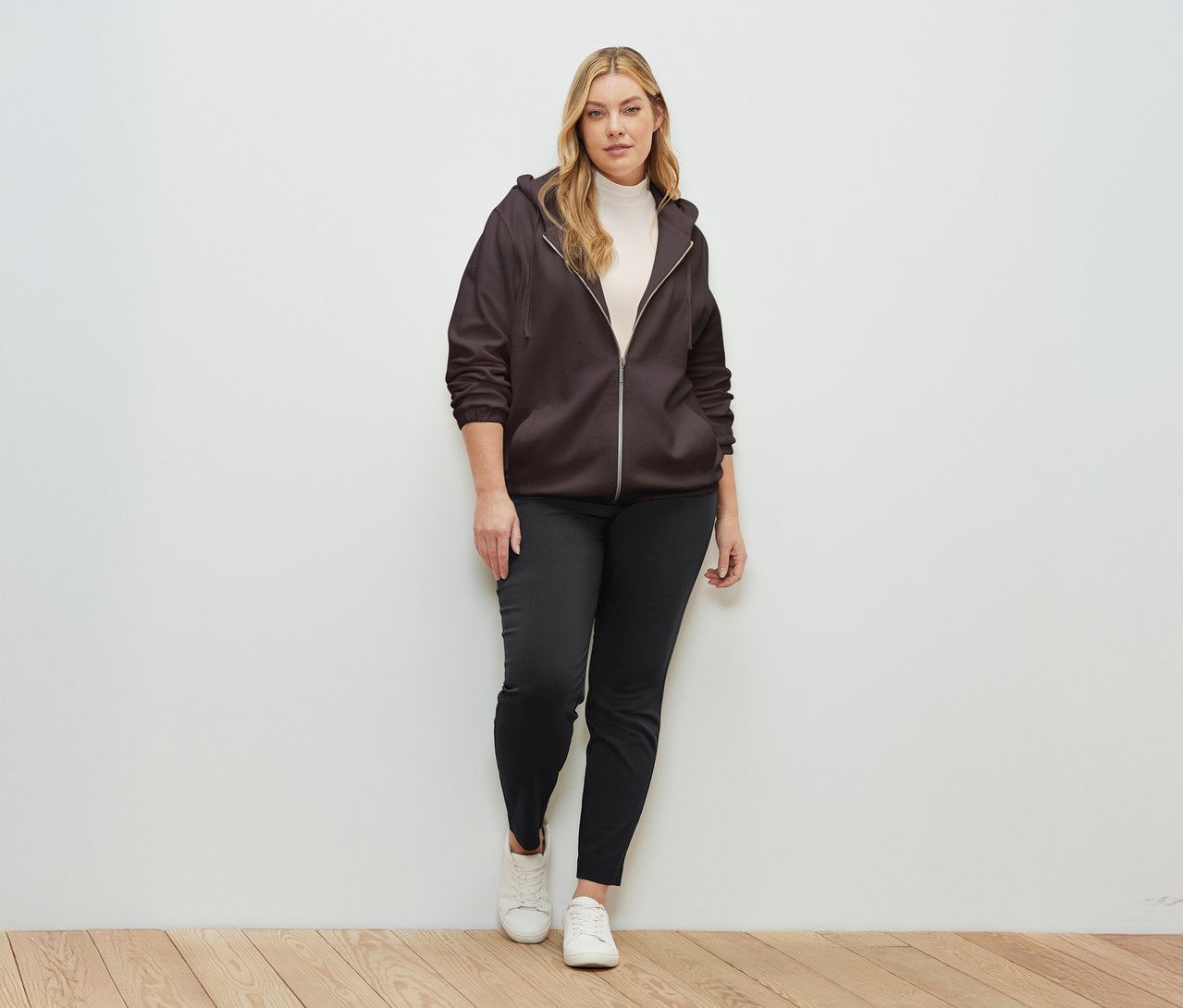 Une femme debout devant un mur portant une veste de survêtement marron foncé.