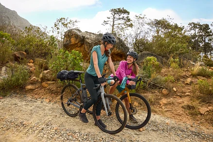 Deux femmes portant des casques se tiennent avec leurs vélos sur un chemin de gravier dans un terrain vallonné. Une gourde est attachée au vélo.