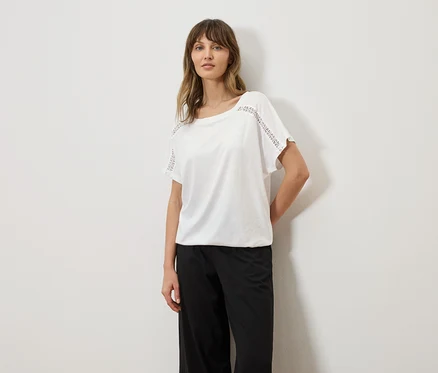 Femme portant un t-shirt blanc avec des détails en broderie anglaise sur les manches et un pantalon noir.