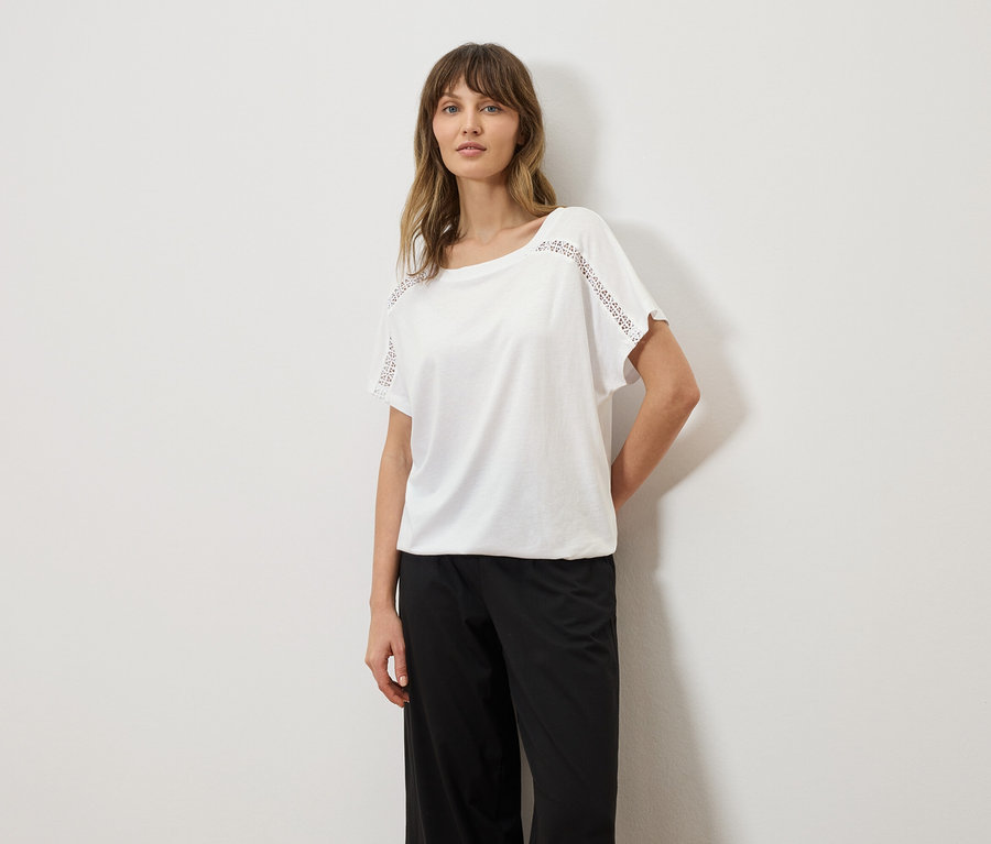 Femme portant un t-shirt blanc avec des détails en broderie anglaise sur les manches et un pantalon noir.