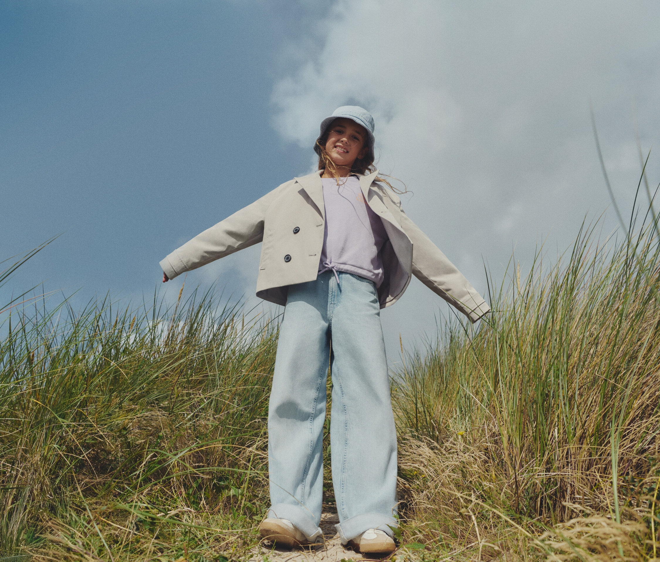 Une jeune fille se tient dans un champ avec de hautes herbes, portant une veste claire, un jean large et un chapeau assorti.