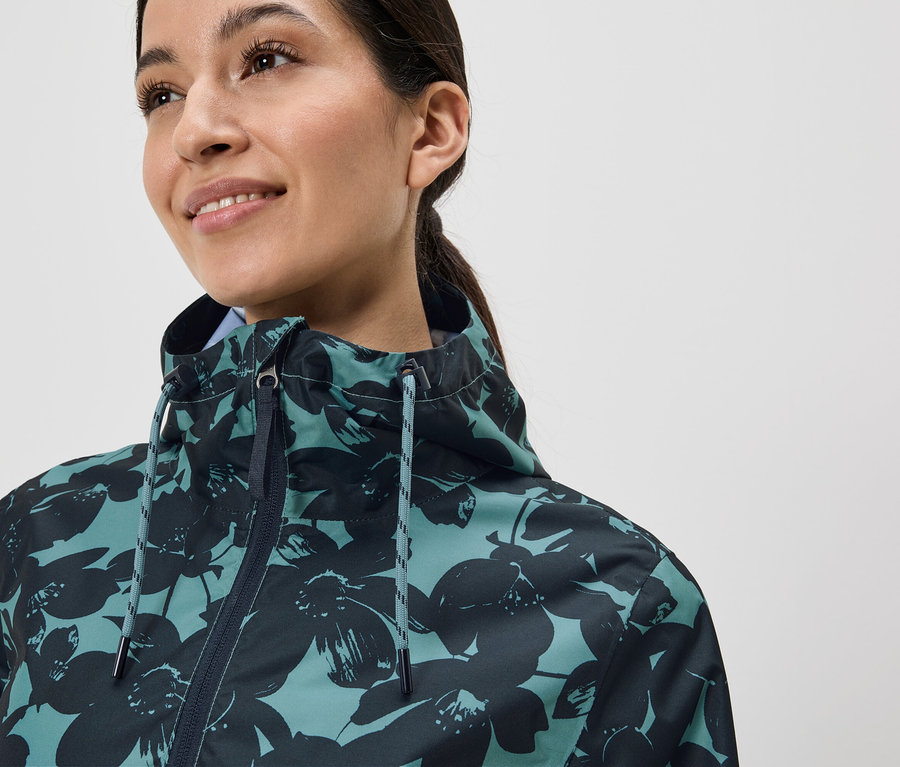 Une femme souriante avec une queue de cheval porte une veste turquoise à fleurs foncées.