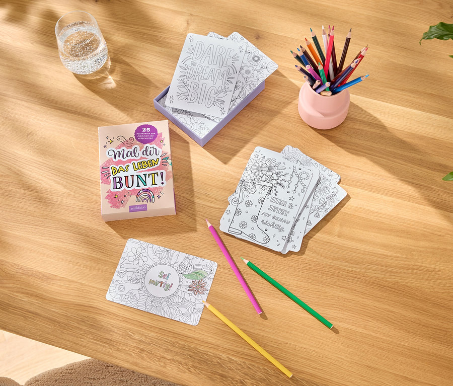Sur une table, il y a des livres de coloriage, des cartes avec des motifs à colorier, des crayons de couleur dans un pot rose et un verre d'eau.