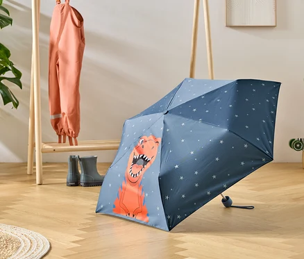 Un parapluie bleu à motif de dinosaure est posé sur un plancher en bois, à côté d'un porte-manteau avec des vêtements de pluie et des bottes en caoutchouc.