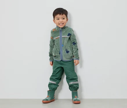 Un garçon souriant se tient devant un mur blanc, vêtu d'une veste en polaire verte à motifs de dinosaures, d'un pantalon vert et de bottes en caoutchouc vertes.