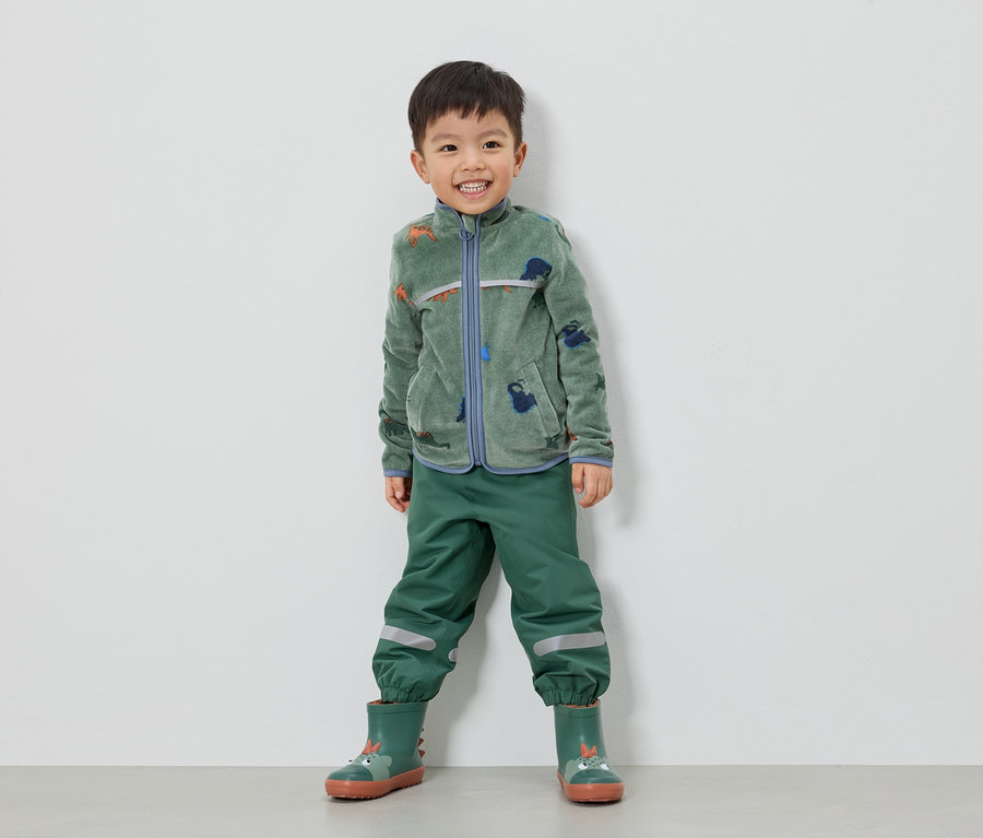 Un garçon souriant se tient devant un mur blanc, vêtu d'une veste en polaire verte à motifs de dinosaures, d'un pantalon vert et de bottes en caoutchouc vertes.