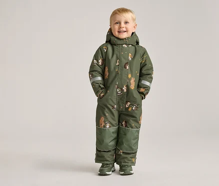 Un garçon blond souriant se tient sur un fond clair, vêtu d'une combinaison verte à motifs d'animaux et de chaussures vertes.