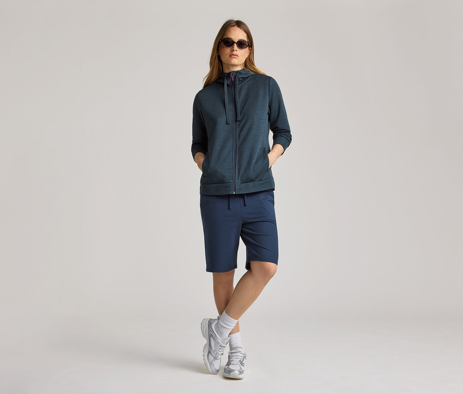 Une femme portant des lunettes de soleil porte un veste à capuche bleu marine et un short. La veste à capuche est ouverte.