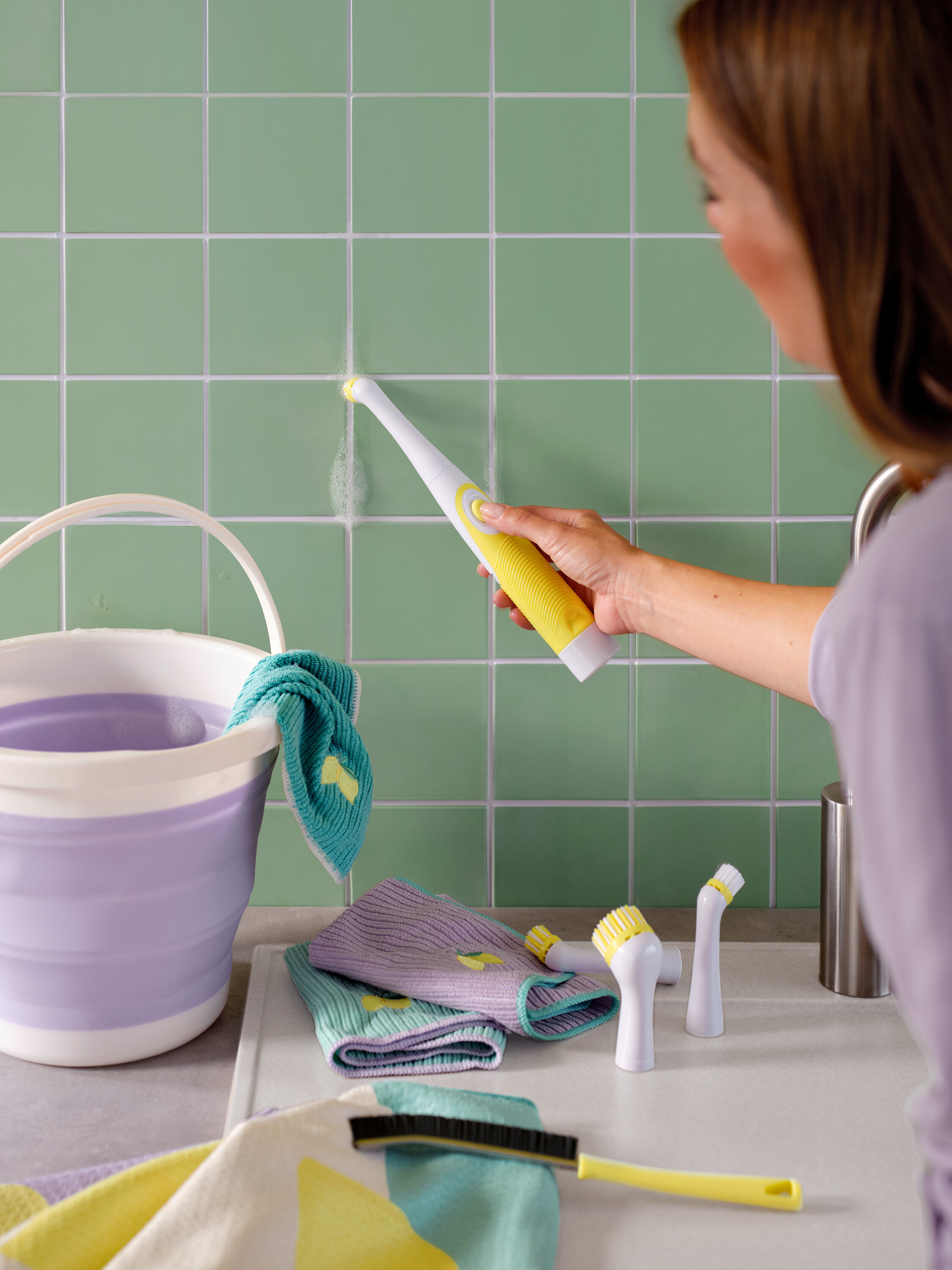 Une femme nettoie des carreaux verts avec une brosse jaune et blanche, un seau et des chiffons sont également visibles.