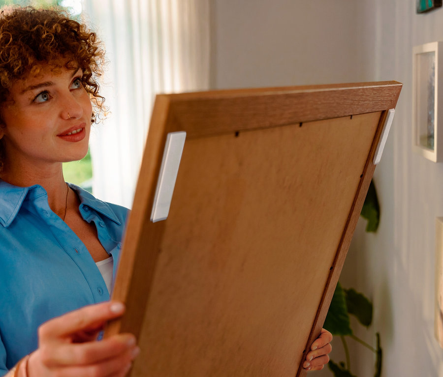 Une femme aux cheveux bouclés tient un cadre en bois avec des bandes adhésives blanches, alors qu'elle l'aligne sur un mur avec d'autres images.