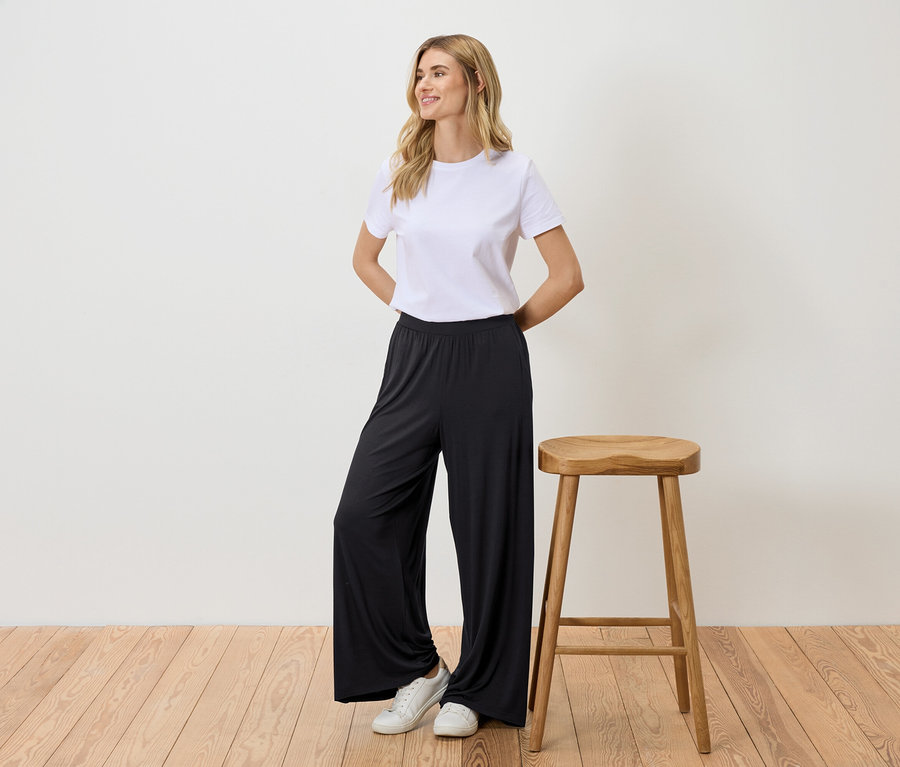 Une femme se tient à côté d'un tabouret, porte un t-shirt blanc, un pantalon noir et des baskets blanches.