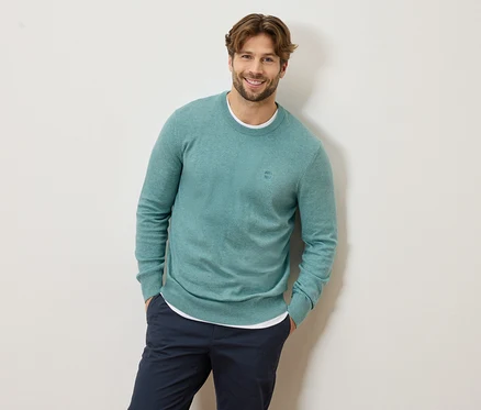 Un homme aux cheveux châtain clair et à la barbe portant un pull bleu clair et un pantalon bleu marine sourit.