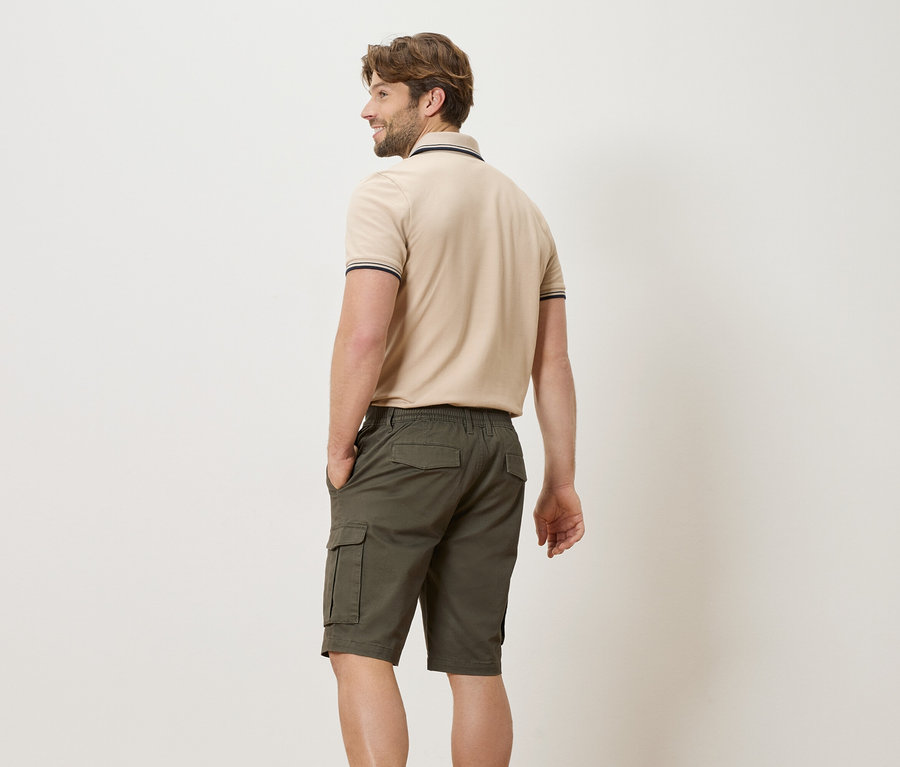 Homme vu de dos portant un polo beige et un short vert avec les mains dans les poches.