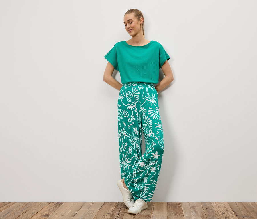 Femme portant un haut vert et un pantalon vert et blanc appuyée contre un mur blanc.