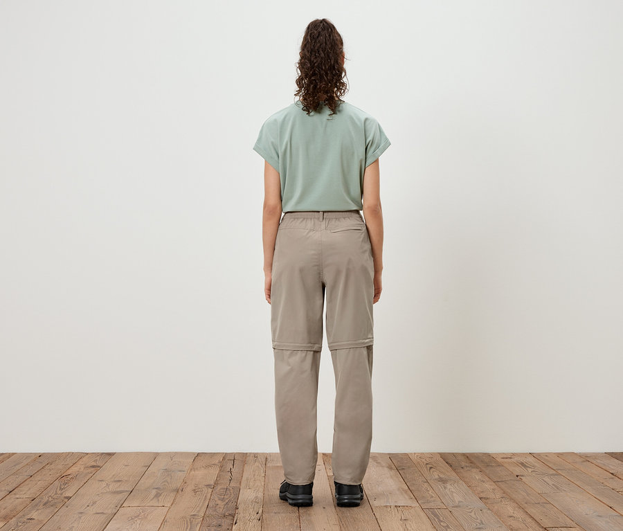 Vue de dos d'une femme aux cheveux ondulés mi-longs portant un t-shirt vert clair, un pantalon convertible marron clair et des chaussures noires.