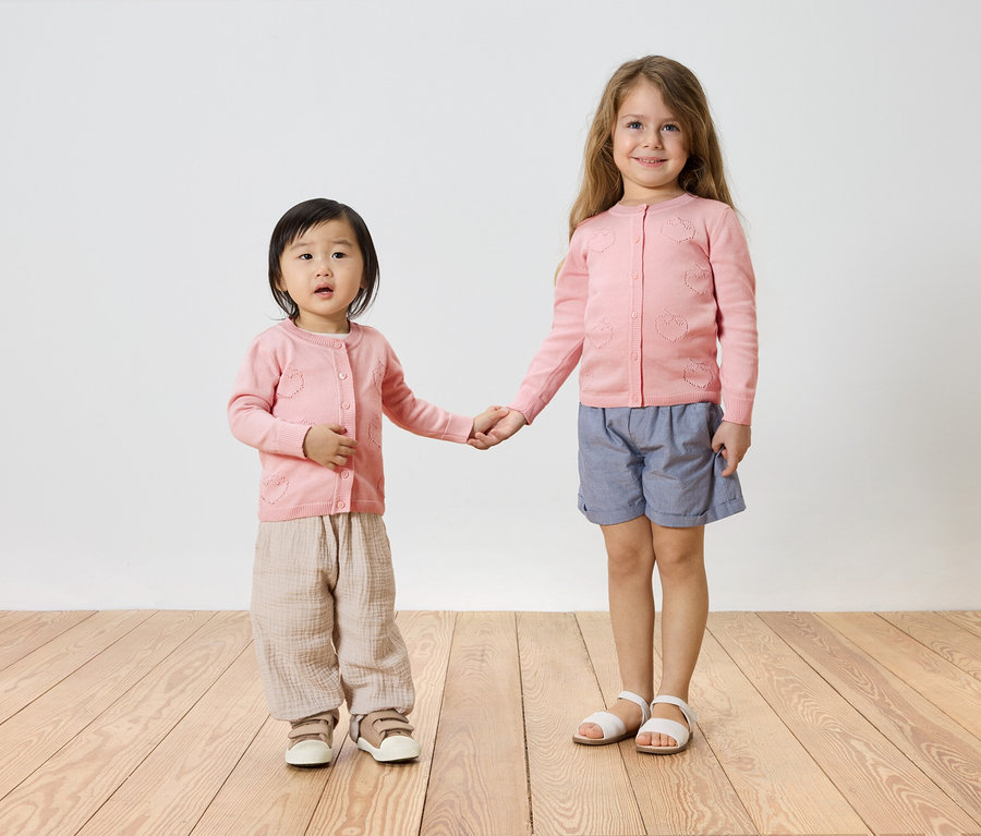 Deux fillettes, une aux cheveux foncés et l'autre aux cheveux blonds, se tiennent la main. Elles portent toutes les deux des gilets roses, la plus petite avec un pantalon beige et des baskets, et la plus grande avec un short bleu et des sandales.