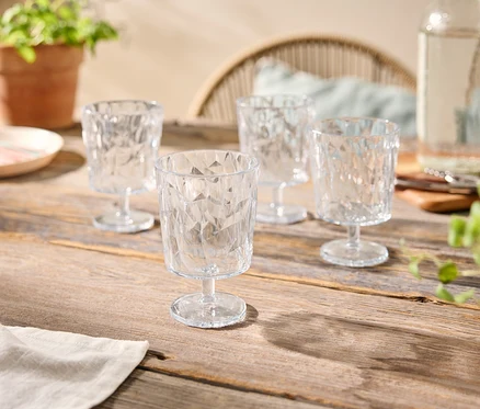 Quatre verres transparents à facettes sur une table en bois, avec une bouteille d'eau, une assiette et des plantes en pot.