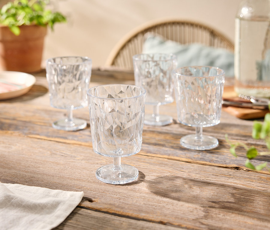 Quatre verres transparents à facettes sur une table en bois, avec une bouteille d'eau, une assiette et des plantes en pot.