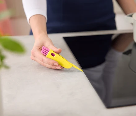 Une femme nettoie une plaque de cuisson noire avec un outil de nettoyage jaune et rose.