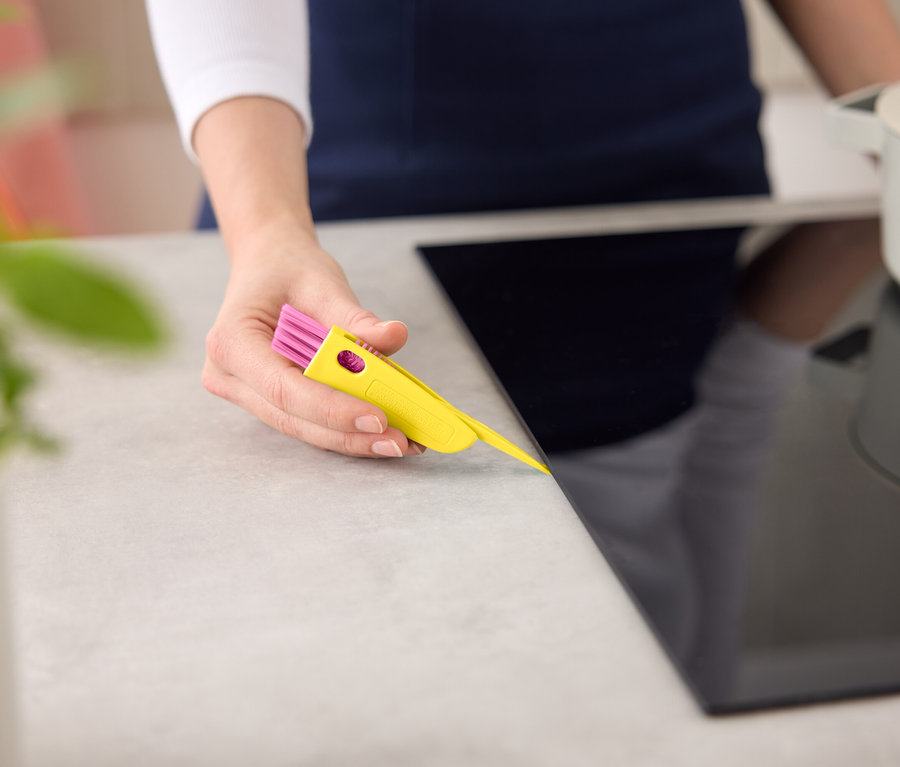 Une femme nettoie une plaque de cuisson noire avec un outil de nettoyage jaune et rose.