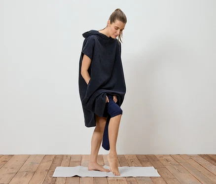 Une femme en poncho de bain bleu marine arrivant à mi-cuisse se tient pieds nus sur un tapis de yoga.