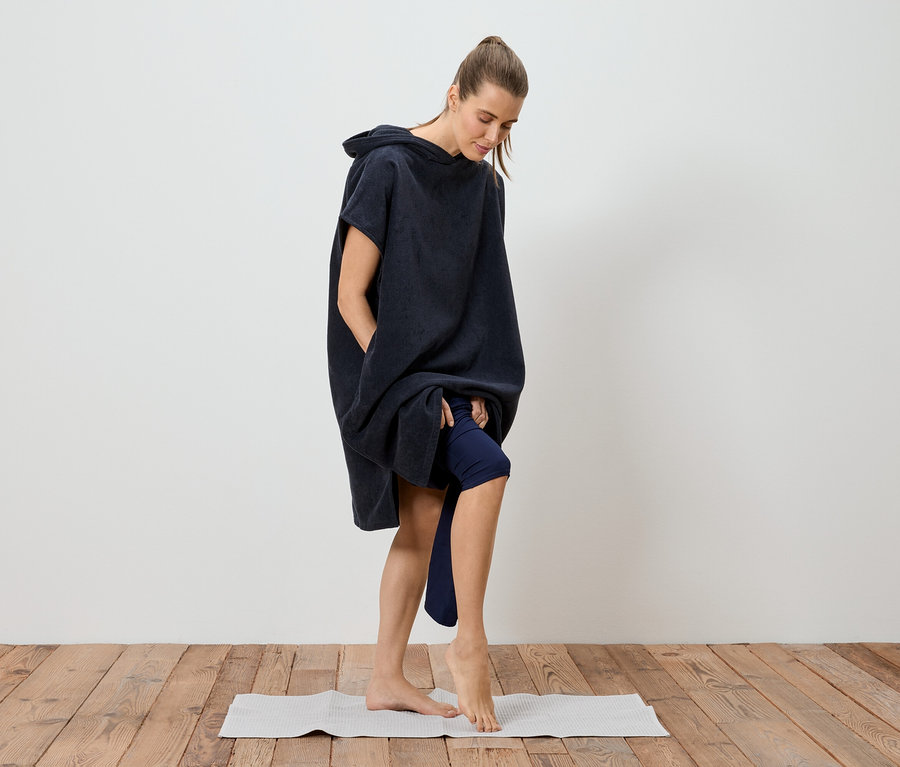 Une femme en poncho de bain bleu marine arrivant à mi-cuisse se tient pieds nus sur un tapis de yoga.