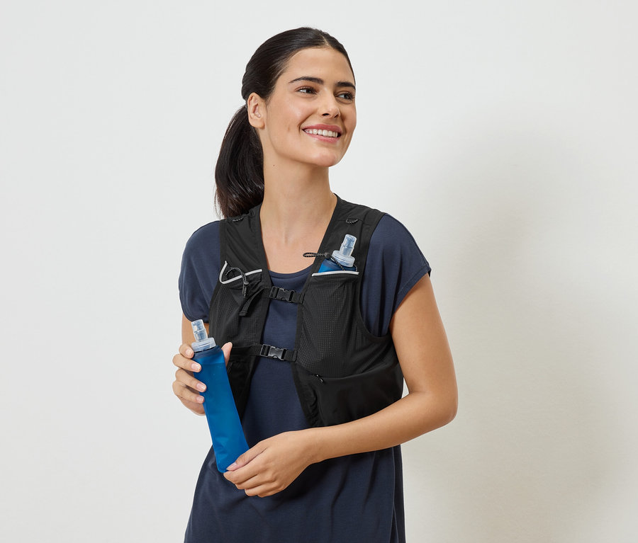 Femme souriante portant un gilet de course noir et tenant une gourde bleue.