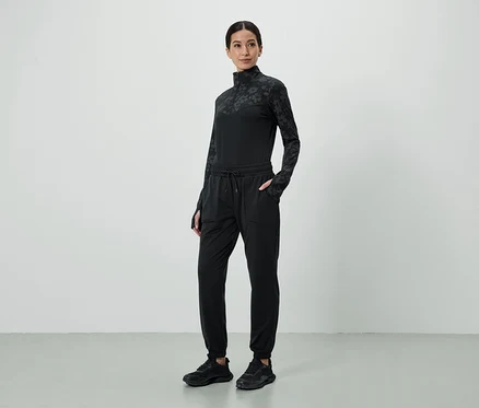 Femme portant un haut noir à motifs et un pantalon de jogging avec les mains dans les poches, ainsi que des baskets noires.