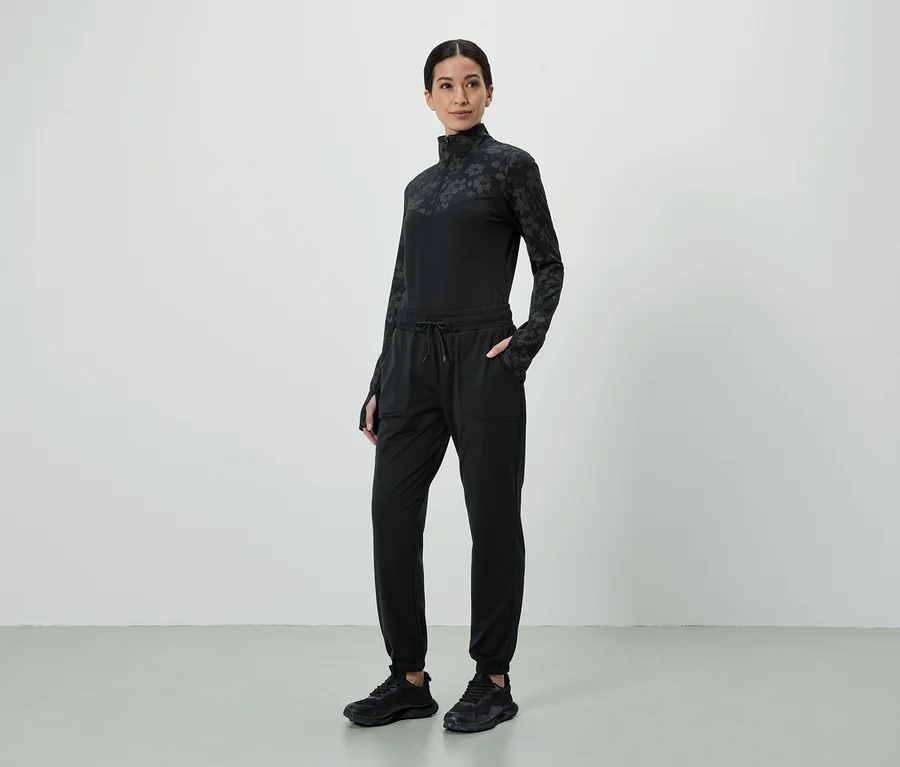 Femme portant un haut noir à motifs et un pantalon de jogging avec les mains dans les poches, ainsi que des baskets noires.