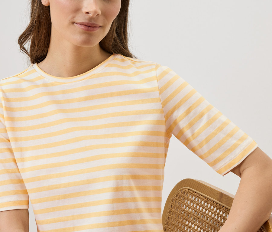 Une femme porte un t-shirt rayé jaune et blanc et tient une chaise en rotin.