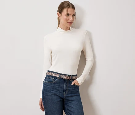 Une femme portant un pull à col roulé crème, un jean et une ceinture à motif animalier se tient devant un mur blanc.