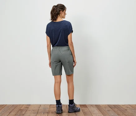 Vue arrière d'une femme portant un t-shirt foncé, un short gris et des chaussures de randonnée devant un mur blanc.