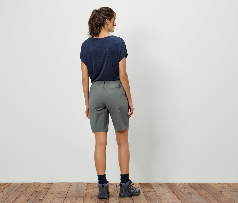 Vue arrière d'une femme portant un t-shirt foncé, un short gris et des chaussures de randonnée devant un mur blanc.