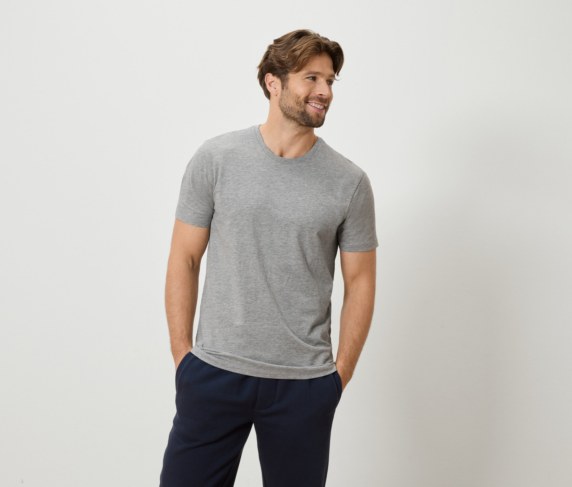 Homme portant un t-shirt gris et un pantalon bleu marine, les mains dans les poches, sur un fond blanc.