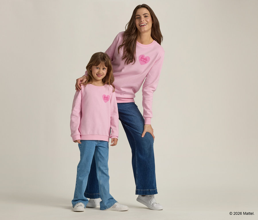 Une femme et une fille se tiennent ensemble et portent des pulls roses avec un logo Barbie en forme de cœur et des jeans.
