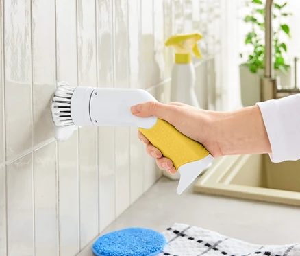 Une main tient une brosse de nettoyage électrique avec une poignée jaune et une tête de brosse blanche, nettoyant les carreaux.