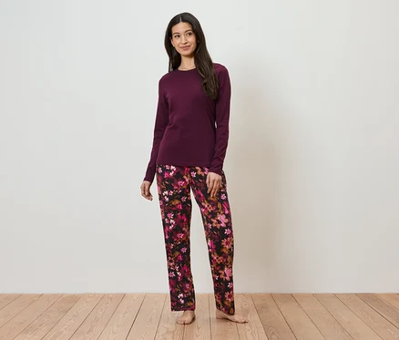 Une femme portant un haut bordeaux et un pantalon à fleurs se tient sur un plancher en bois devant un mur blanc.